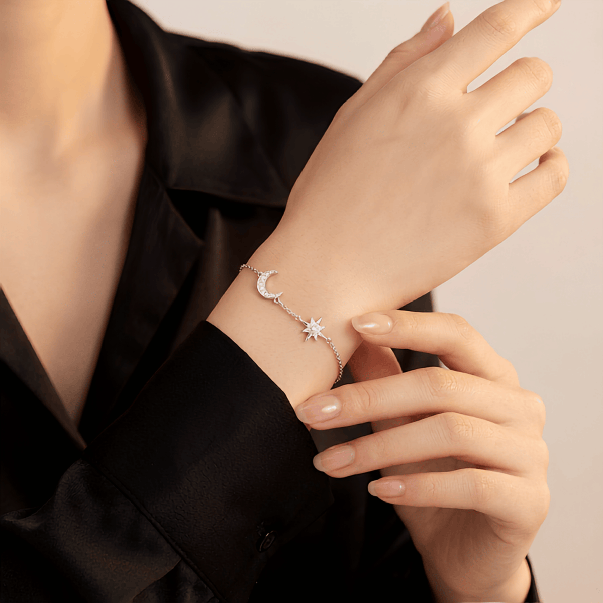 A delicate silver bracelet with a crescent moon and star design, shown being worn on a person's wrist. The person is wearing a black satin blazer against a soft, light beige background.