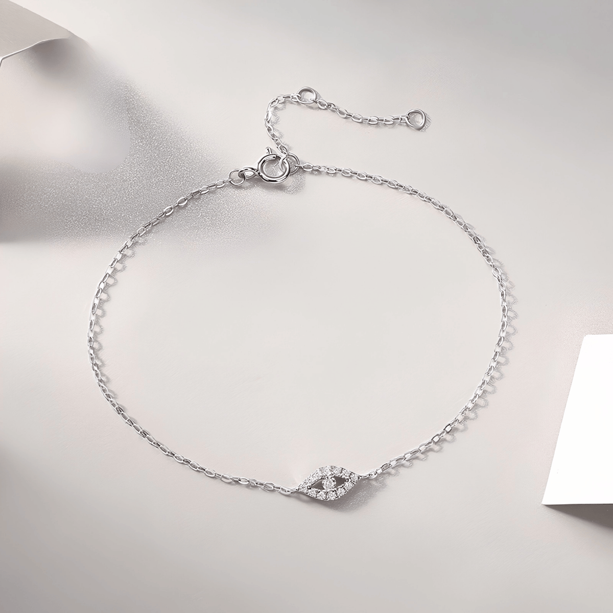 A delicate silver chain bracelet with a small pavé-set eye charm, displayed on a light gray textured surface.
