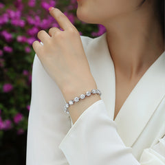 A close-up lifestyle shot of a woman wearing a sparkling silver-toned diamond bracelet on her wrist, dressed in a white blouse with a blurred background of pink flowers.