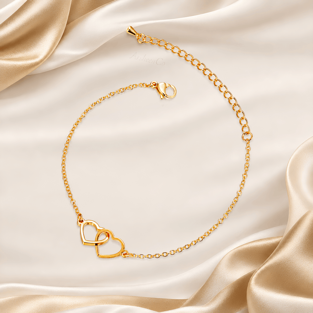 A delicate gold chain anklet featuring a double interlocking heart charm, displayed on draped champagne-colored satin fabric.
