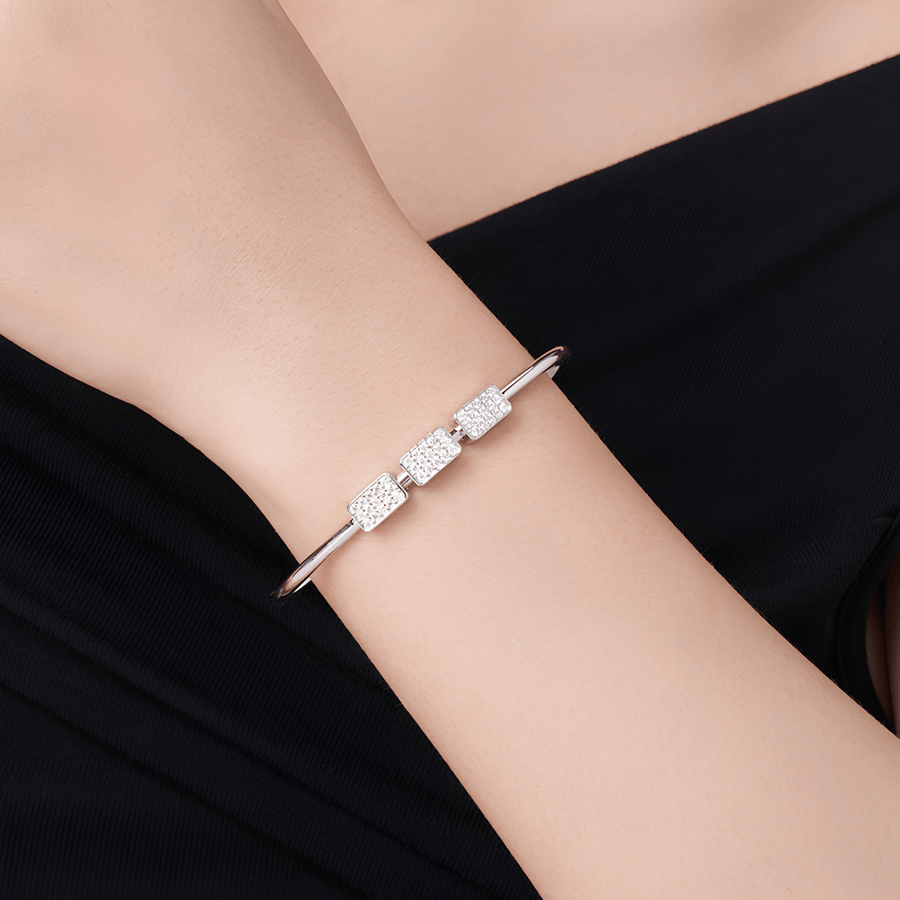 A close-up of a person's wrist wearing a slim silver cuff bracelet with three rectangular bezel-set clear stones, against a black top background.