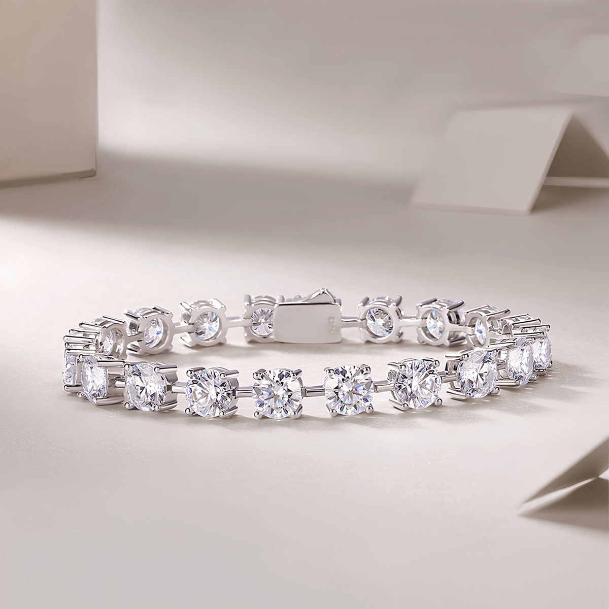A silver tennis bracelet with round-cut clear gemstones, displayed on a clean, light beige surface with soft shadows.