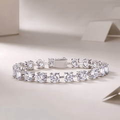 A silver tennis bracelet with round-cut clear gemstones, displayed on a clean, light beige surface with soft shadows.