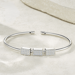 A polished silver-tone open cuff bracelet with three rectangular bezel-set clear stones in the center, displayed on a light textured surface.