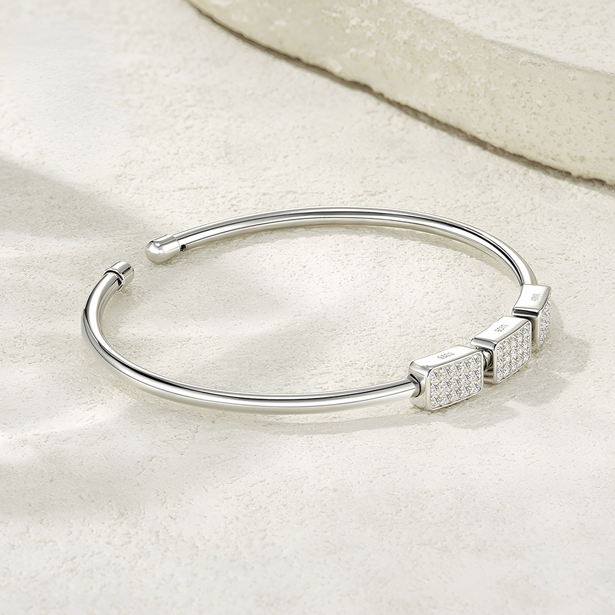 A polished silver open cuff bangle bracelet with three square bezel-set crystal accents, displayed on a textured beige surface.
