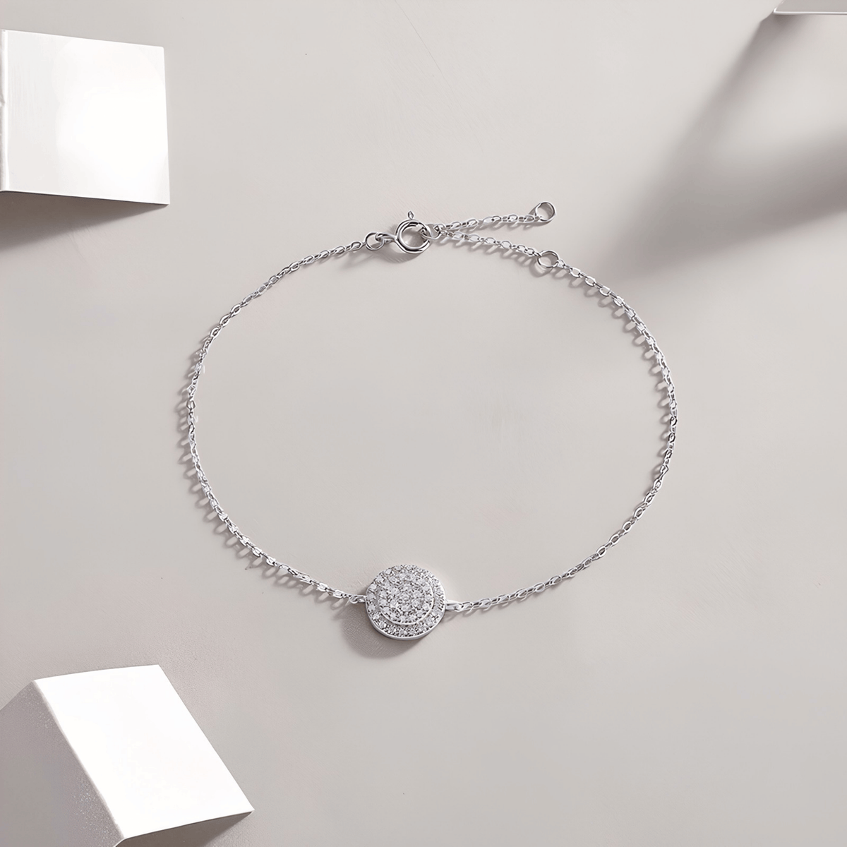 A delicate silver chain bracelet with a round pavé-set stone centerpiece, displayed on a light beige background with minimalist white geometric props.