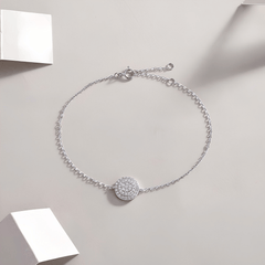 A delicate silver chain bracelet with a round pavé-set stone centerpiece, displayed on a light beige background with minimalist white geometric props.