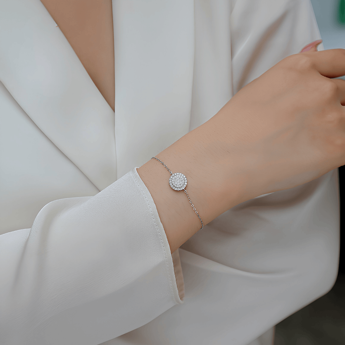 A delicate silver chain bracelet with a round pavé disc charm worn on a woman's wrist, shown in a close-up lifestyle shot with a white satin blouse.