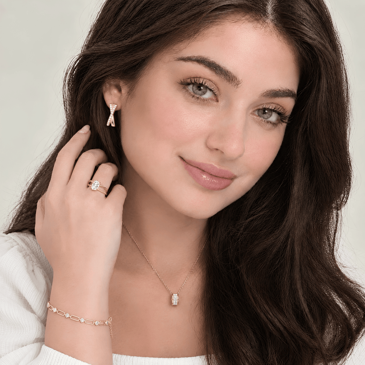 A close-up lifestyle shot of a woman with long dark brown hair wearing a matching gold jewelry set, including a bow-shaped stud earring, a delicate necklace with a small rectangular pendant, and a bracelet with clear stones.