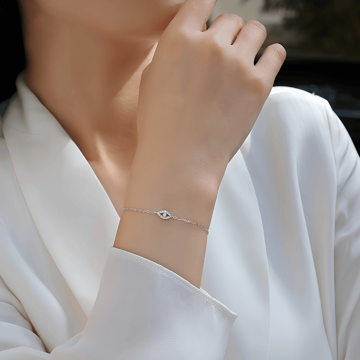 A delicate silver bracelet with a small evil eye charm worn on a woman's wrist, shown in a close-up lifestyle shot against a white satin blouse.