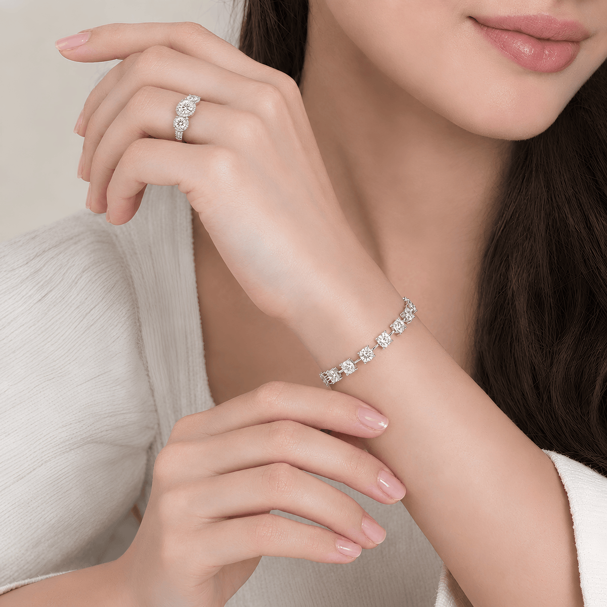 A close-up lifestyle shot of a woman wearing a delicate diamond bracelet on her wrist and a matching diamond ring on her finger, dressed in a soft white top against a warm beige background.