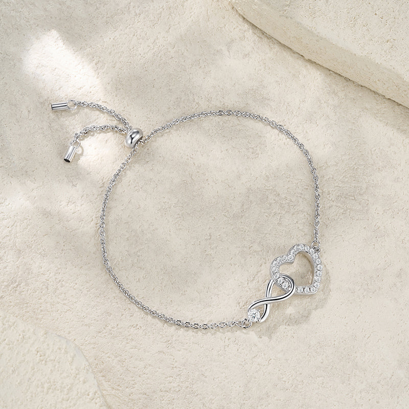A delicate silver chain bracelet with an infinity heart design paved with small clear stones, resting on a textured beige stone surface.