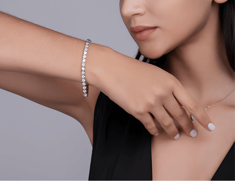 A close-up lifestyle shot of a model wearing a delicate silver tennis bracelet on her wrist, paired with a matching silver necklace on her neck. The model is wearing a black top against a solid gray studio background.