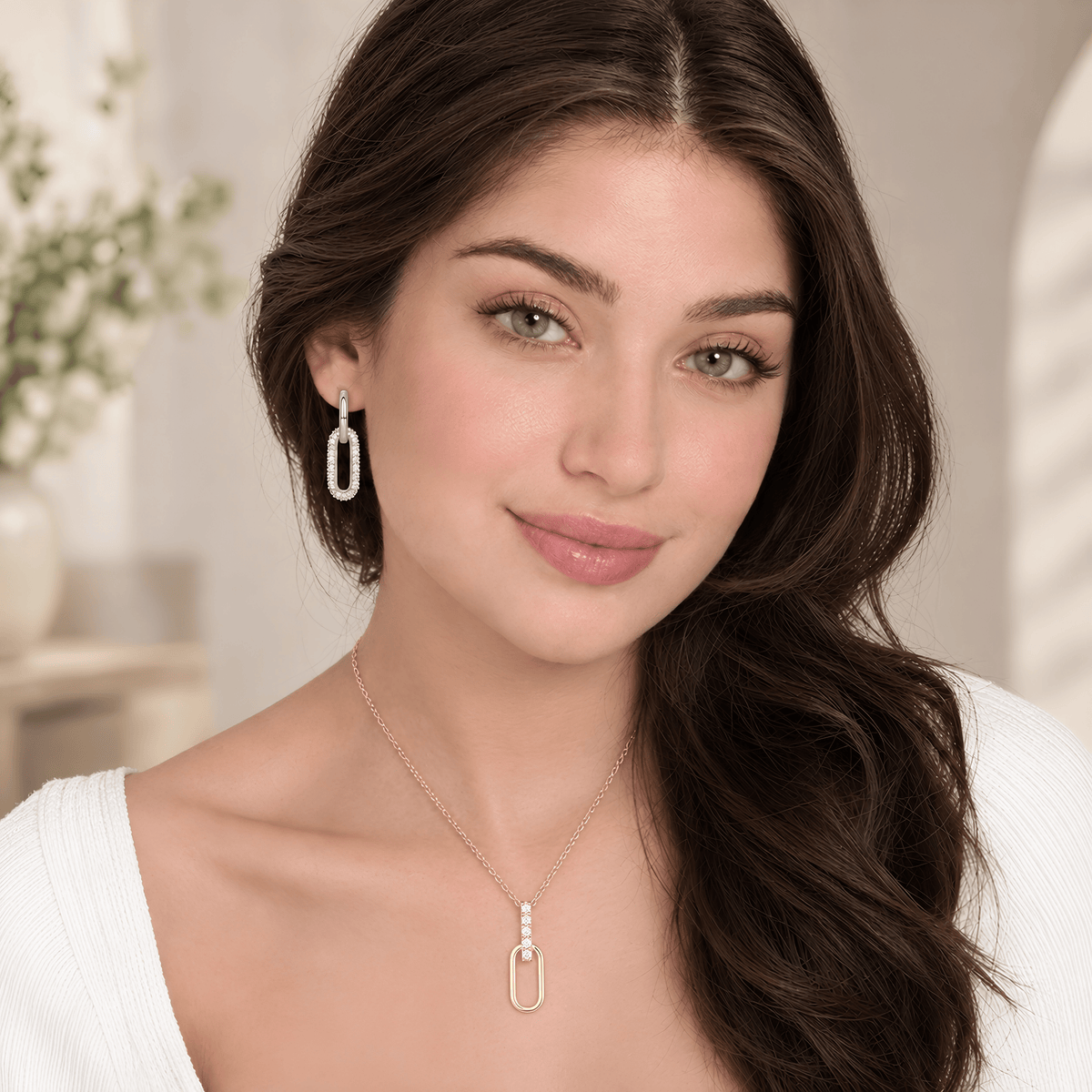 A woman with long dark brown hair models a delicate gold chain necklace with a matching elongated oval pendant and a matching drop earring, set against a softly blurred, warm-toned indoor background.