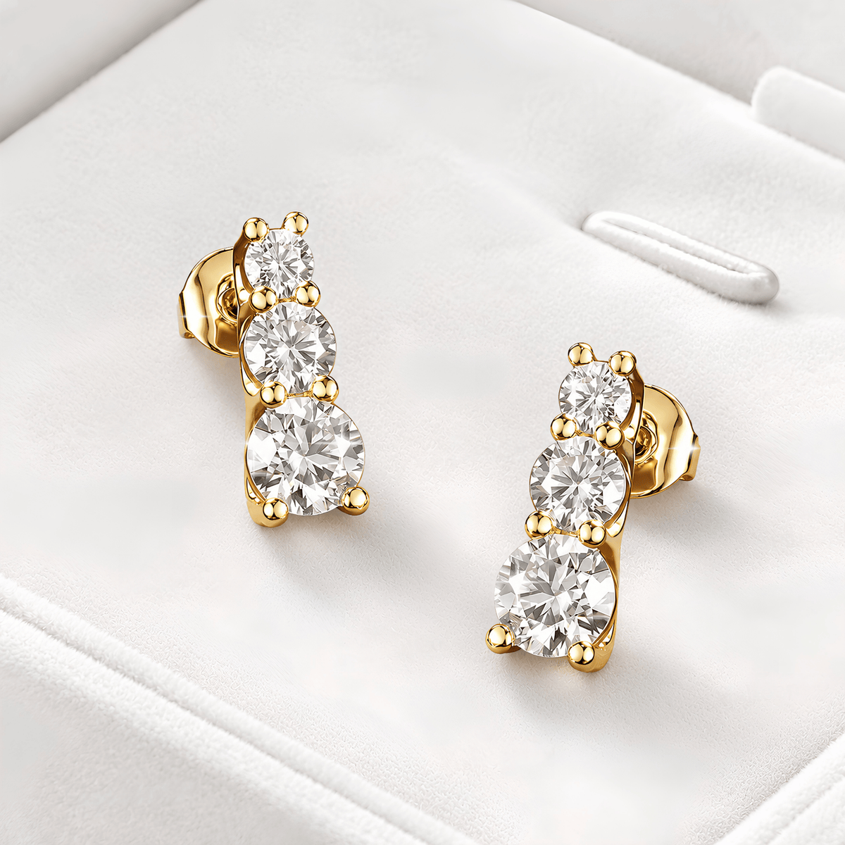 A pair of gold post earrings with three round clear stones on each earring, displayed on a white jewelry box.