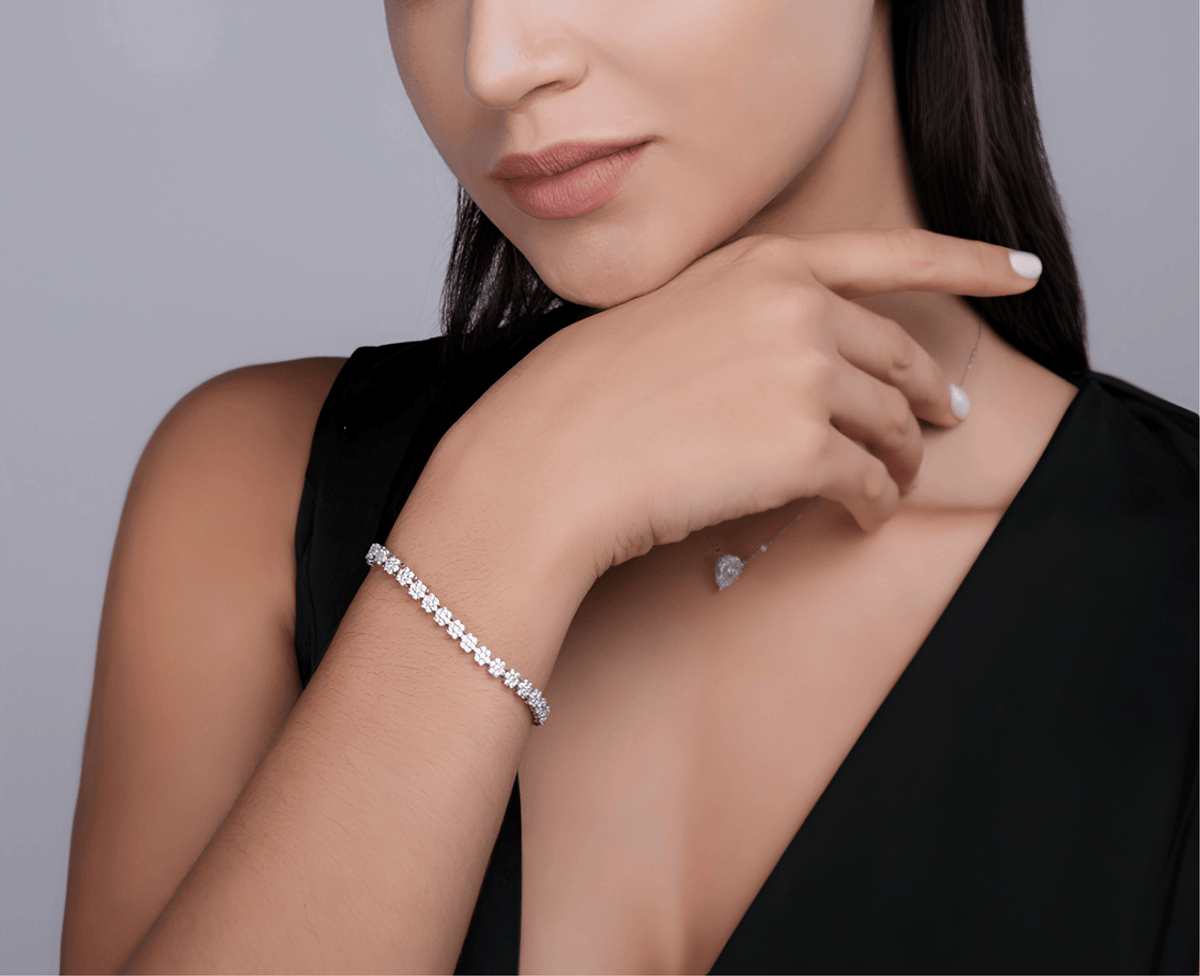 A close-up lifestyle shot of a woman wearing a sparkling silver tennis bracelet on her wrist, paired with a delicate silver necklace. She is wearing a black top against a neutral gray background.