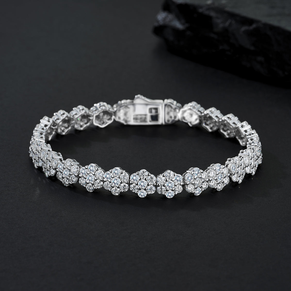 A silver-toned tennis bracelet adorned with round-cut clear stones in a floral cluster setting, displayed on a dark textured surface.