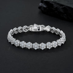 A silver-toned tennis bracelet adorned with round-cut clear stones in a floral cluster setting, displayed on a dark textured surface.