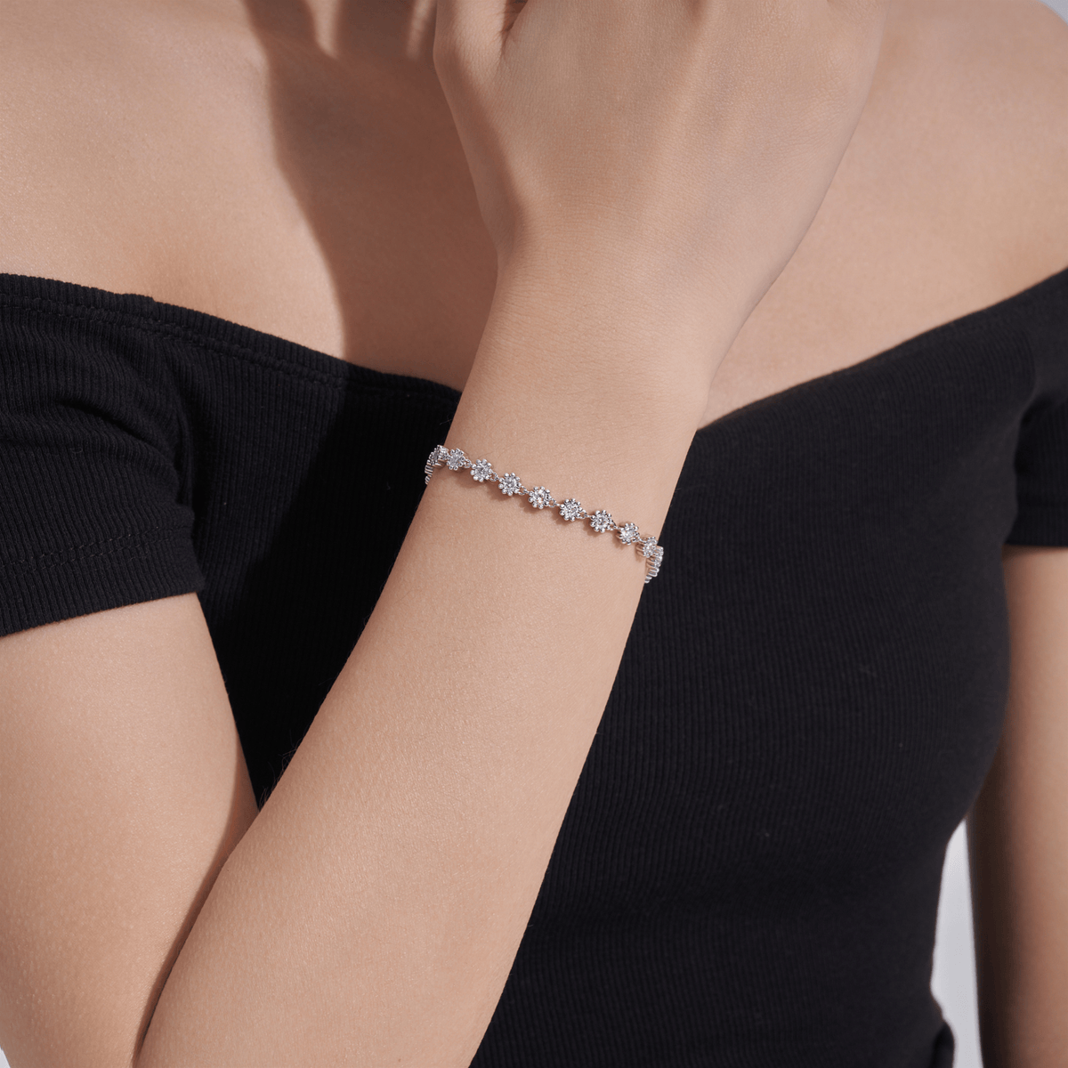 A delicate silver-toned bracelet with small clear gemstones, worn on a woman's wrist. The model is wearing a black off-the-shoulder top, and the background is a clean white studio setting.