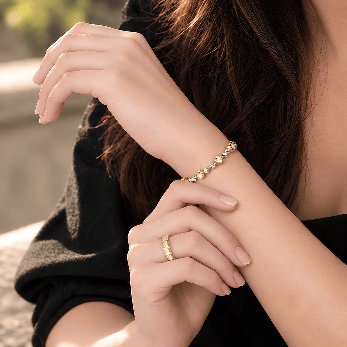 A close-up lifestyle shot of a model wearing a delicate gold and silver beaded bracelet on her wrist, paired with a matching gold ring on her finger. She is wearing a black knit top, and the background is a softly blurred outdoor setting.