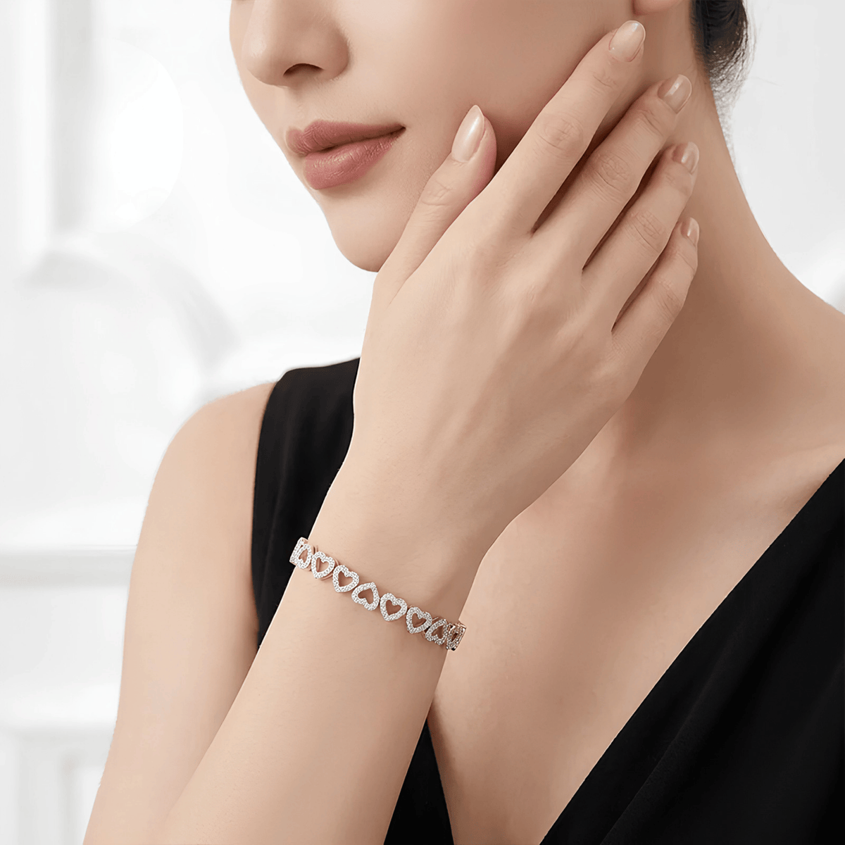 A close-up lifestyle shot of a woman wearing a delicate silver heart-link bracelet on her wrist, with her hand resting near her face against a bright, softly blurred background.