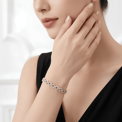 A close-up lifestyle shot of a woman wearing a delicate silver heart-link bracelet on her wrist, with her hand resting near her face against a bright, softly blurred background.