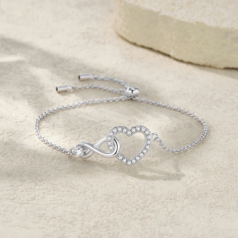 A silver chain bracelet with an infinity symbol and a heart-shaped outline set with small clear stones, displayed on a light beige textured surface.