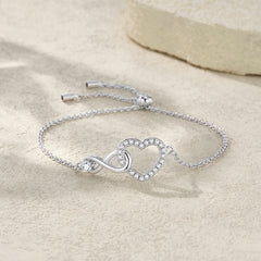 A silver chain bracelet with an infinity symbol and a heart-shaped outline set with small clear stones, displayed on a light beige textured surface.