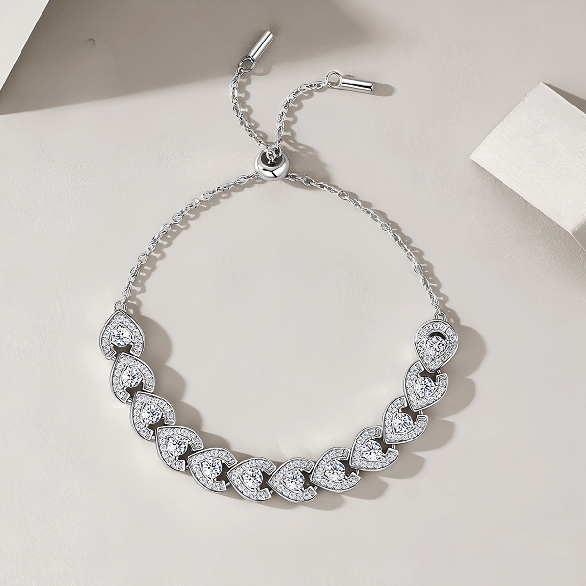 A silver-toned chain bracelet with a row of heart-shaped pavé gemstone clusters, displayed on a light beige surface with soft shadows.