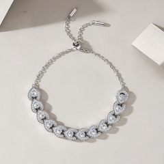 A silver-toned chain bracelet with a row of heart-shaped pavé gemstone clusters, displayed on a light beige surface with soft shadows.