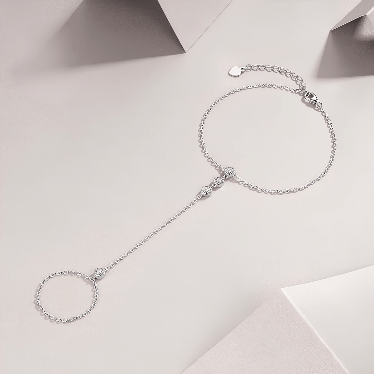 A delicate silver chain anklet with a small heart charm, a central bead detail, and a long dangling chain ending in a circular ring, displayed on a light beige background.