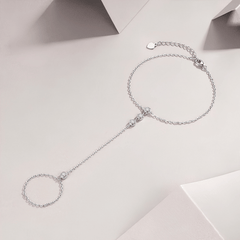 A delicate silver chain anklet with a small heart charm, a central bead detail, and a long dangling chain ending in a circular ring, displayed on a light beige background.