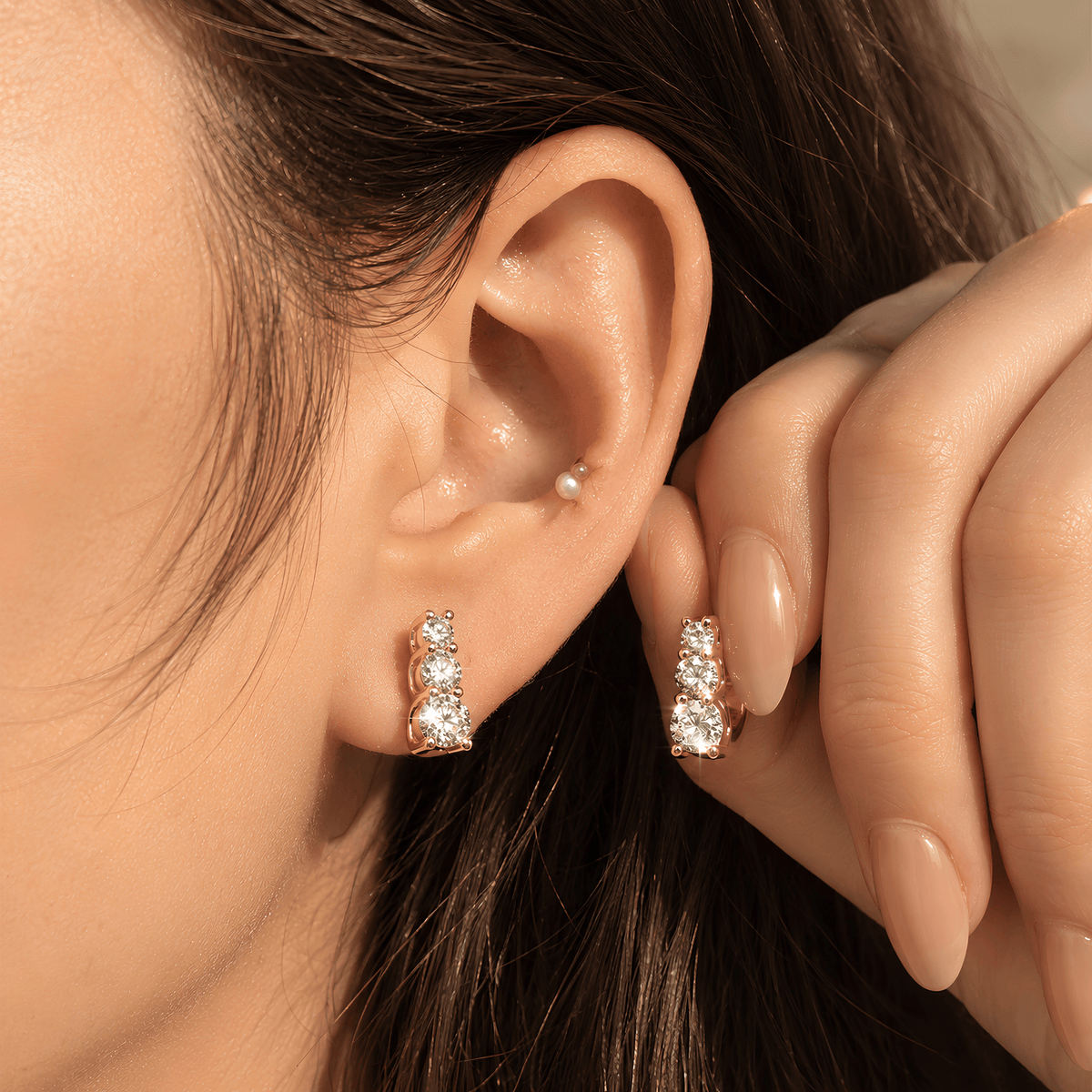 Close-up of a person's ear wearing a rose gold huggie hoop earring with three clear stones, alongside a matching pearl stud in the lobe and a small gold hoop in the cartilage.
