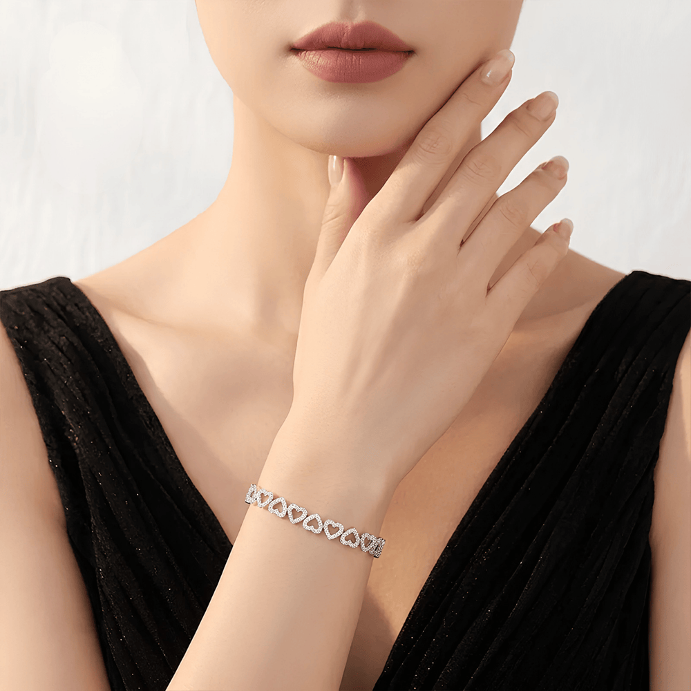 A close-up of a model wearing a delicate silver bracelet with a row of interlocking heart shapes, paired with a black sleeveless top against a soft light gray background.