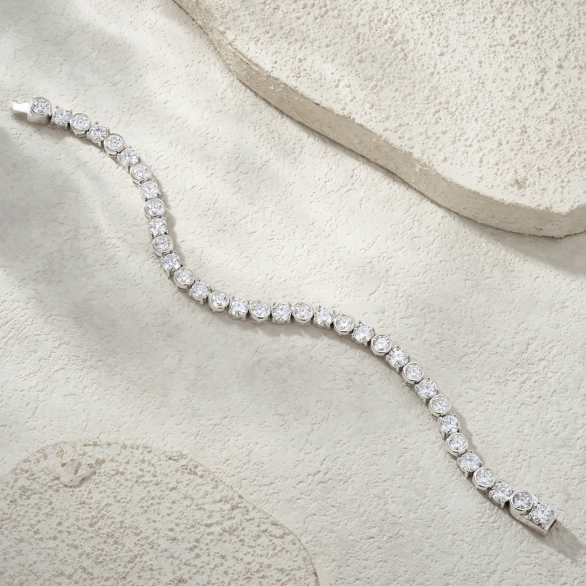 A silver tennis bracelet with round-cut clear stones, arranged in a curved line on a textured beige stone surface.