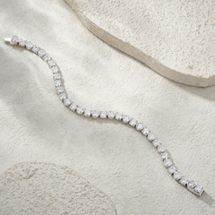 A silver tennis bracelet with round-cut clear stones, arranged in a curved line on a textured beige stone surface.