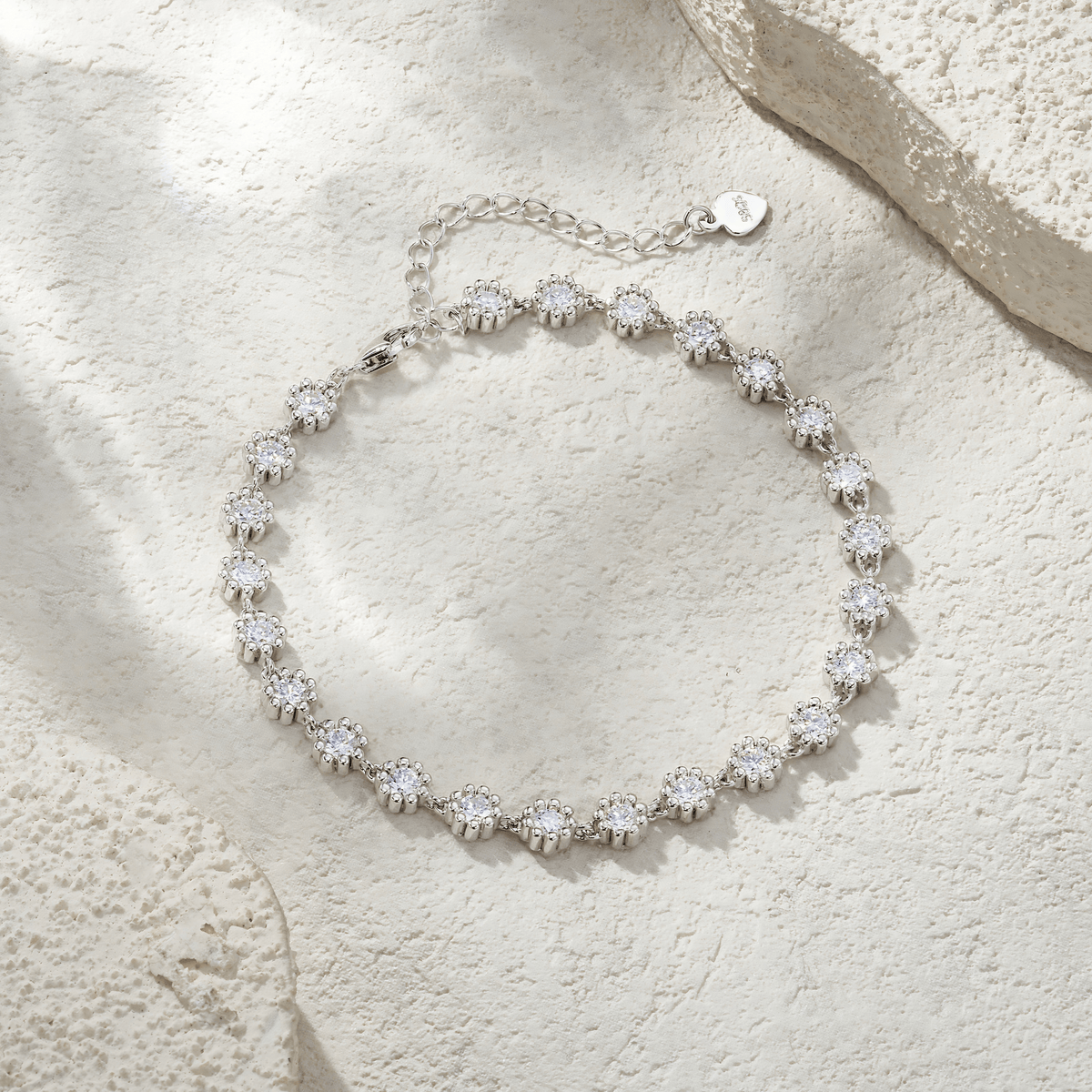 A silver-toned tennis bracelet with round-cut clear stones, arranged in a circle on a textured beige stone surface with soft shadows.