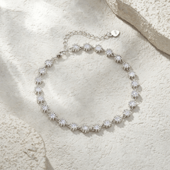 A silver-toned tennis bracelet with round-cut clear stones, arranged in a circle on a textured beige stone surface with soft shadows.