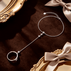 A silver chain anklet with a crescent moon pendant and a dangling ring, displayed on a dark brown velvet surface with ornate gold picture frames and satin bows.