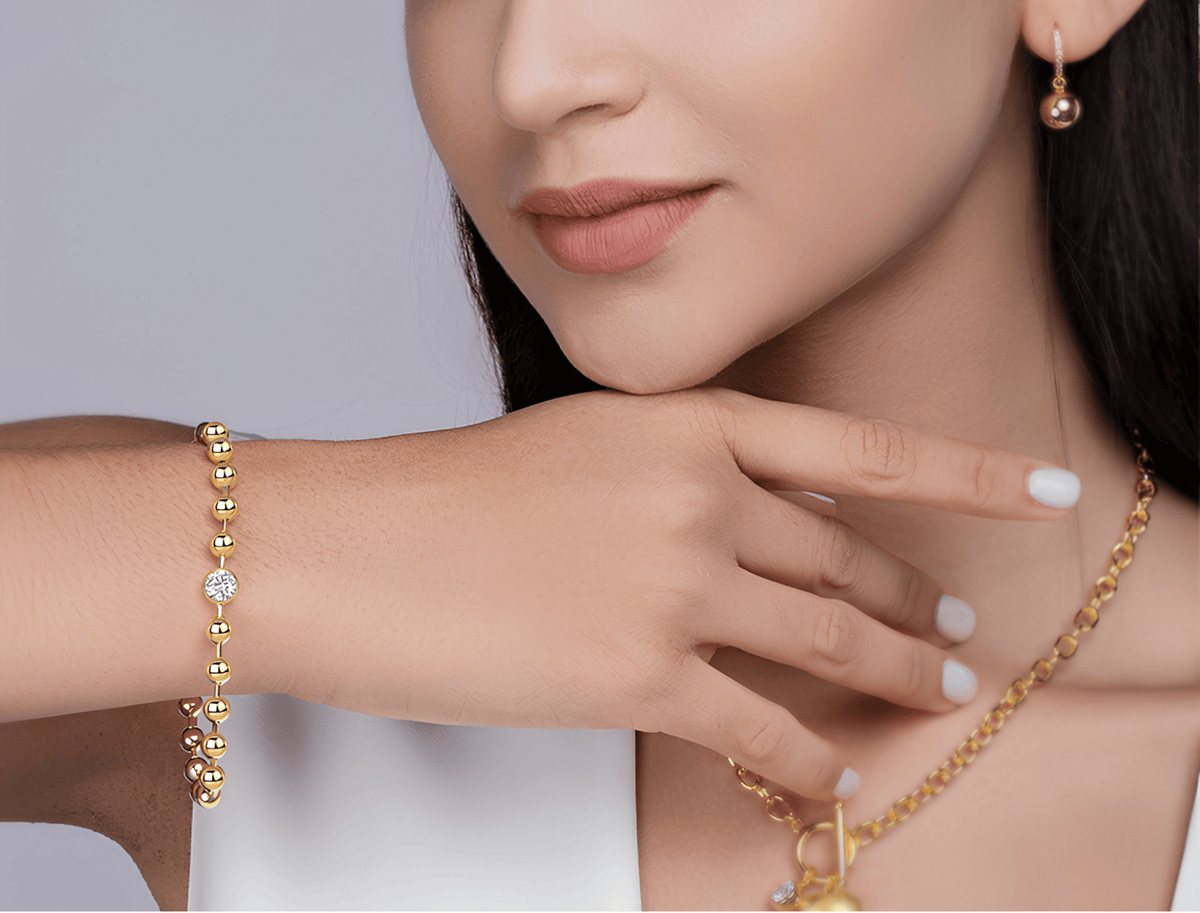 A close-up lifestyle shot of a model wearing a gold beaded bracelet on her wrist, a matching gold chain necklace, and a small gold hoop earring against a soft gray background.