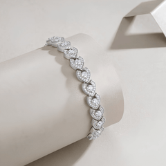 A silver-toned heart-link bracelet with clear pavé stones, displayed on a cream-colored cylindrical jewelry stand against a light beige background.
