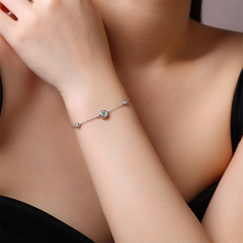 A delicate silver chain bracelet with a central round bezel-set clear stone and small side accents, shown worn on a woman's wrist against a dark top background.