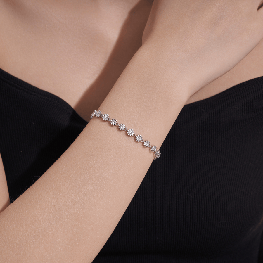 A silver-toned tennis bracelet featuring a continuous row of round clear gemstones in a floral or sunburst setting, with a lobster clasp and an adjustable chain extender with a heart-shaped tag, shown on a plain white background.