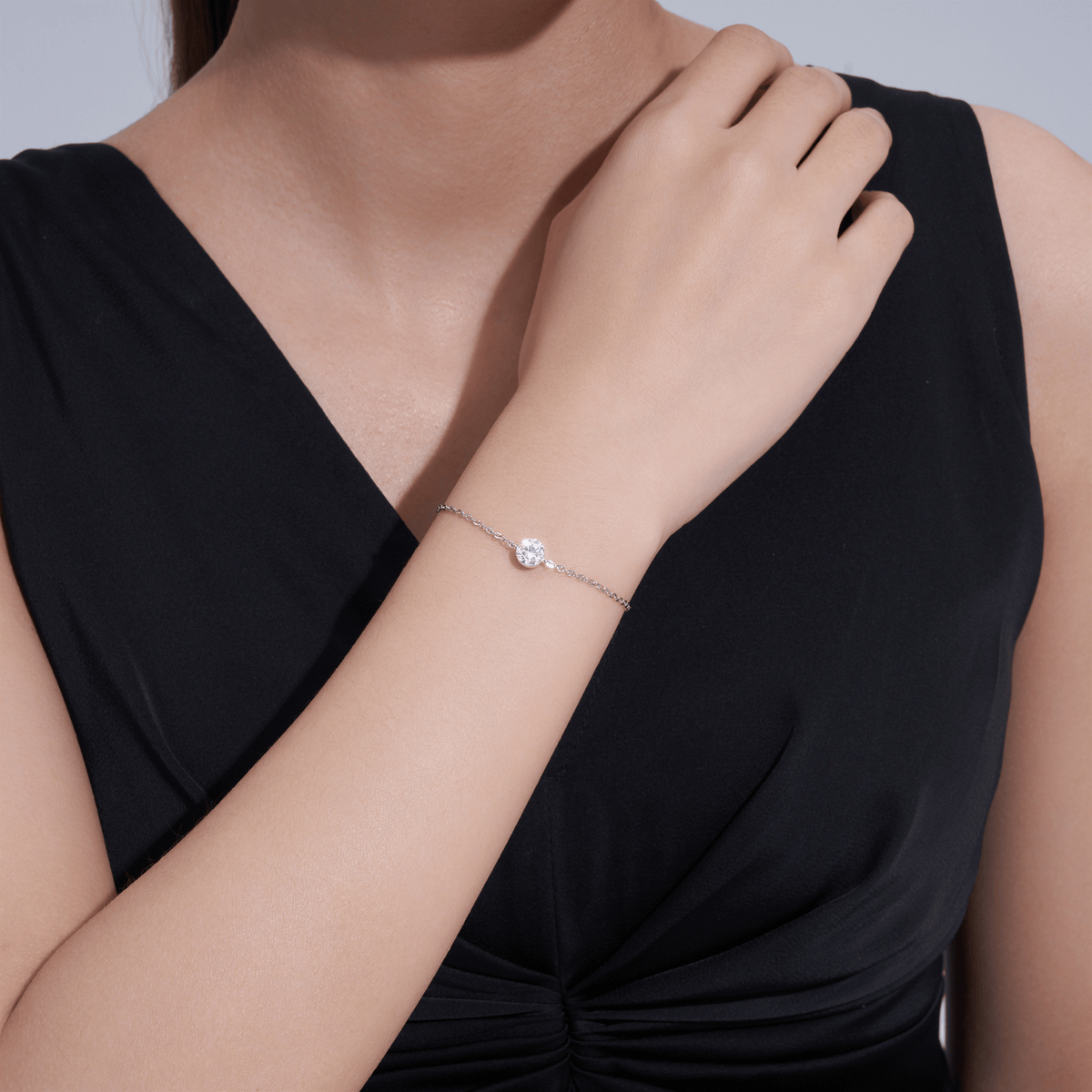 A close-up of a model wearing a delicate silver chain bracelet with a small, clear, flower-like crystal charm. The model is wearing a black sleeveless top, and her hand rests near her neck.