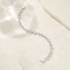 A silver tennis bracelet with a variety of gemstone shapes and sizes, laid out on a textured beige fabric background.