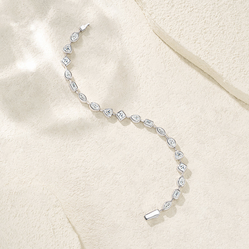 A silver tennis bracelet with a variety of gemstone shapes and sizes, laid out on a textured beige fabric background.