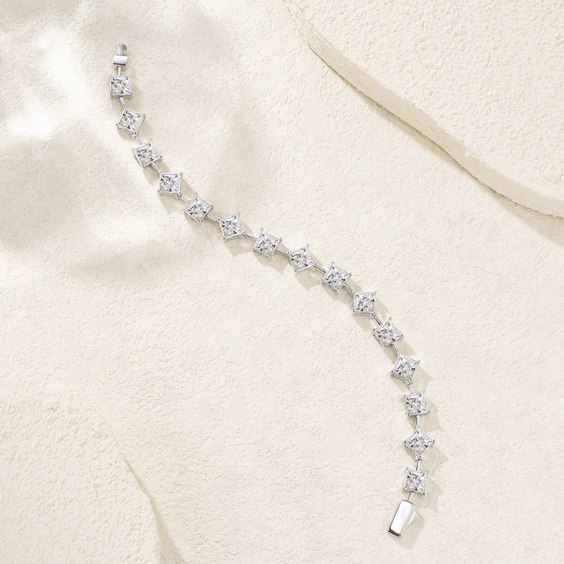 A silver tennis bracelet with clear gemstones, arranged in a curved line on a textured beige fabric background.