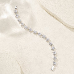 A silver tennis bracelet with clear gemstones, arranged in a curved line on a textured beige fabric background.