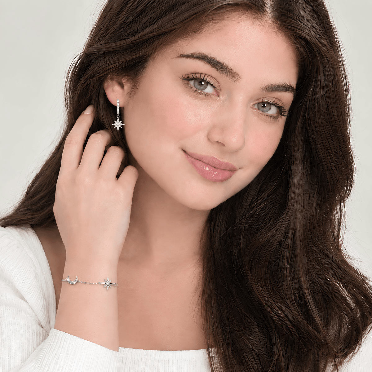 A woman with long dark brown hair models a delicate silver-toned jewelry set, featuring a star-shaped stud earring and a matching crescent moon and star bracelet, against a clean, light off-white background.