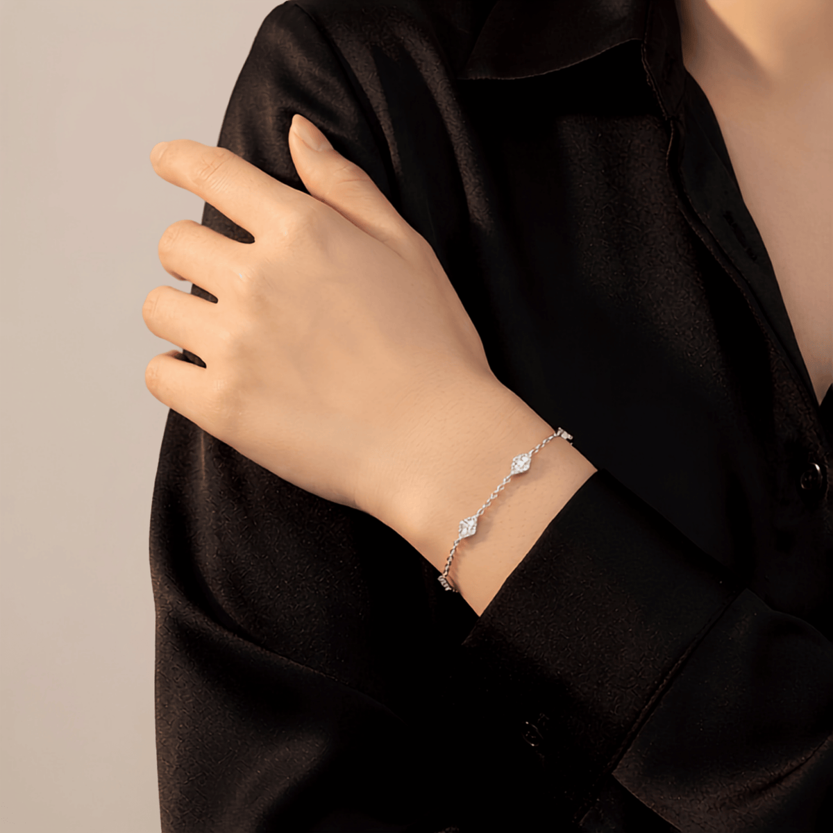 A delicate silver chain bracelet with three small diamond-shaped charms, shown worn on a person's wrist. The person is wearing a black satin button-up shirt against a neutral light gray background.